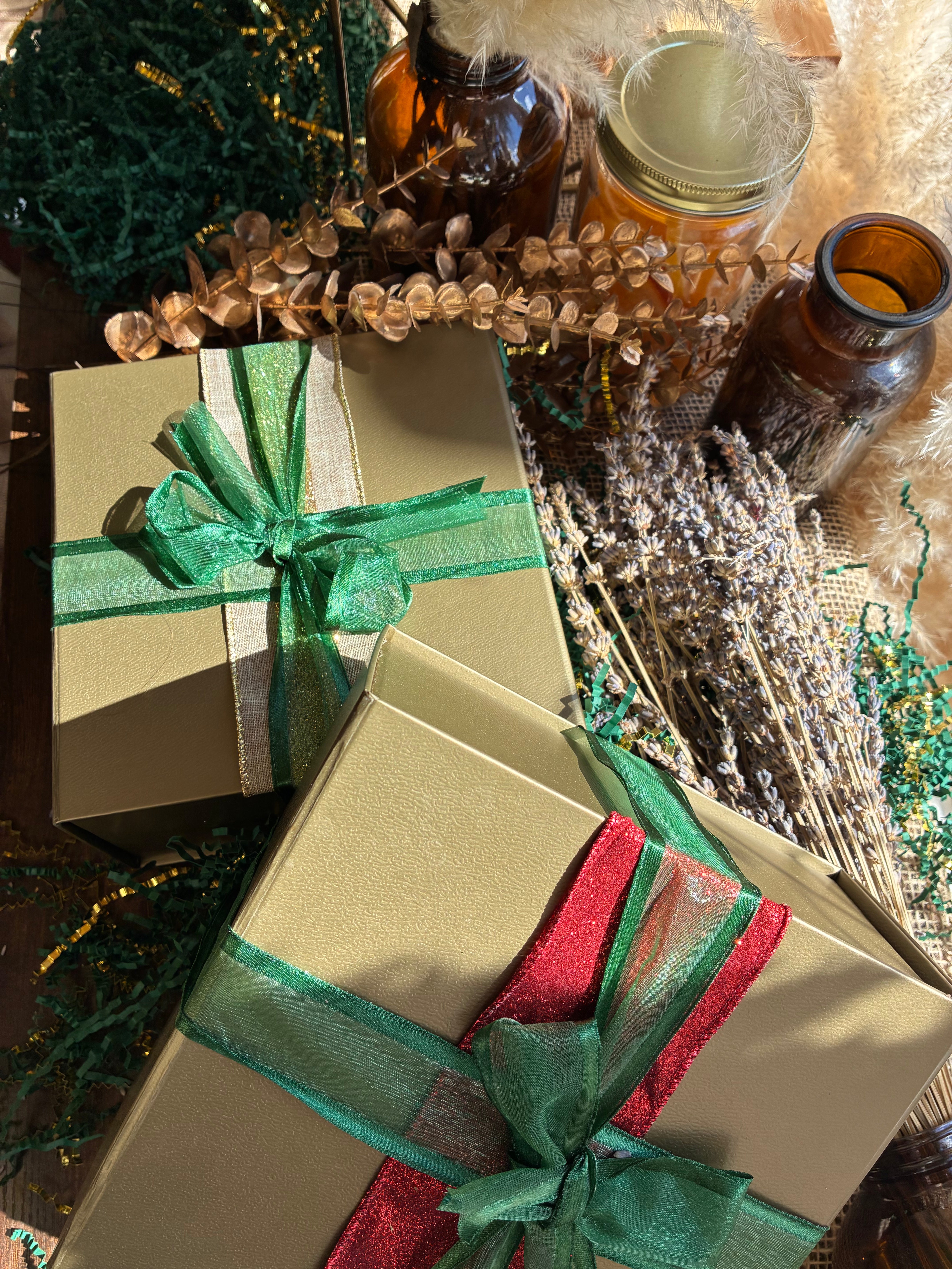Two square kraft gift boxes Holiday wrapped in shiny green and red ribbons, styled with dried flowers, amber glass bottles, and decorative greenery on a tabletop.