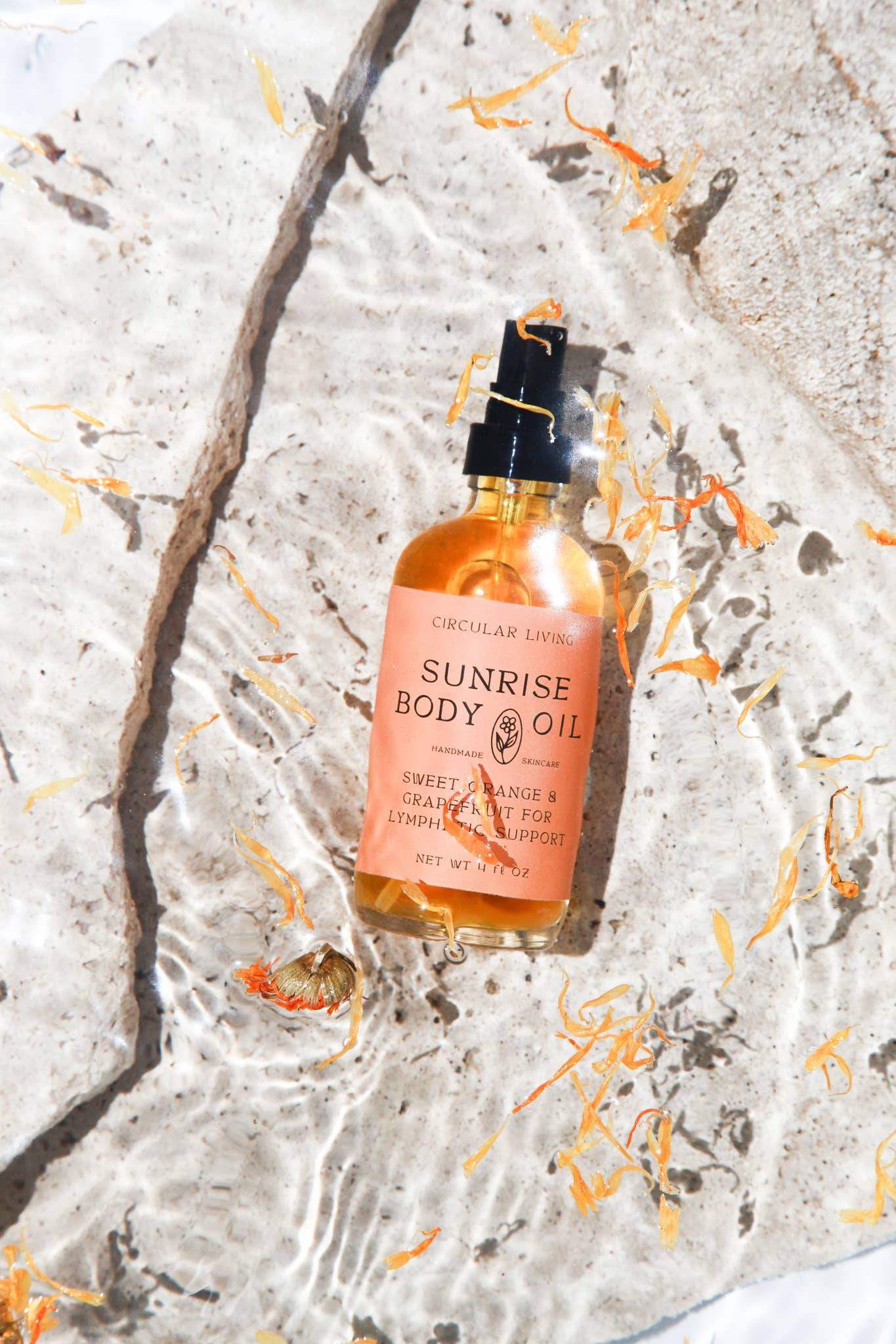 Bottle of Sunrise Body Oil on a textured surface with orange petals
