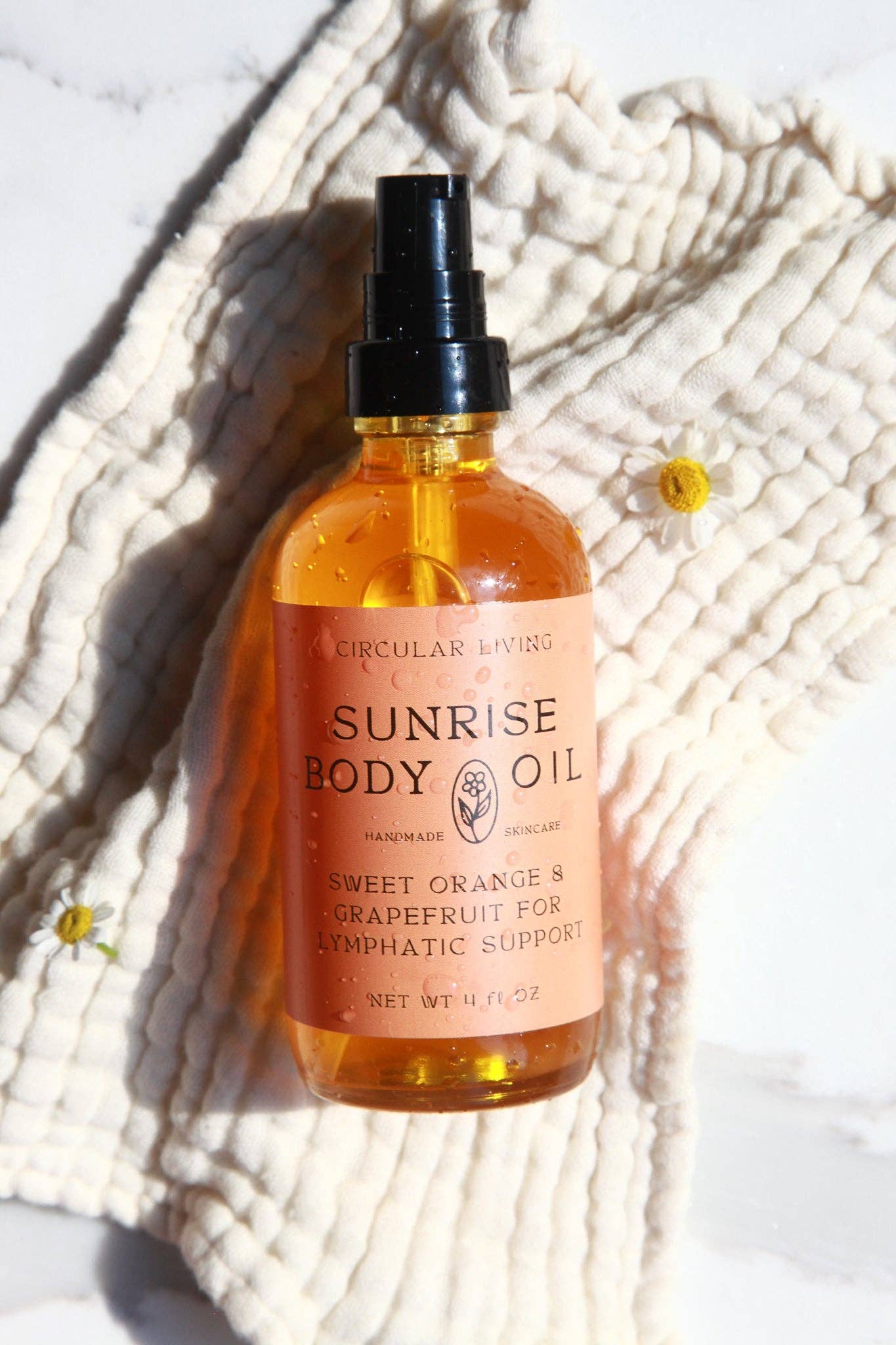 Bottle of Sunrise Body Oil on a textured white surface with flowers.