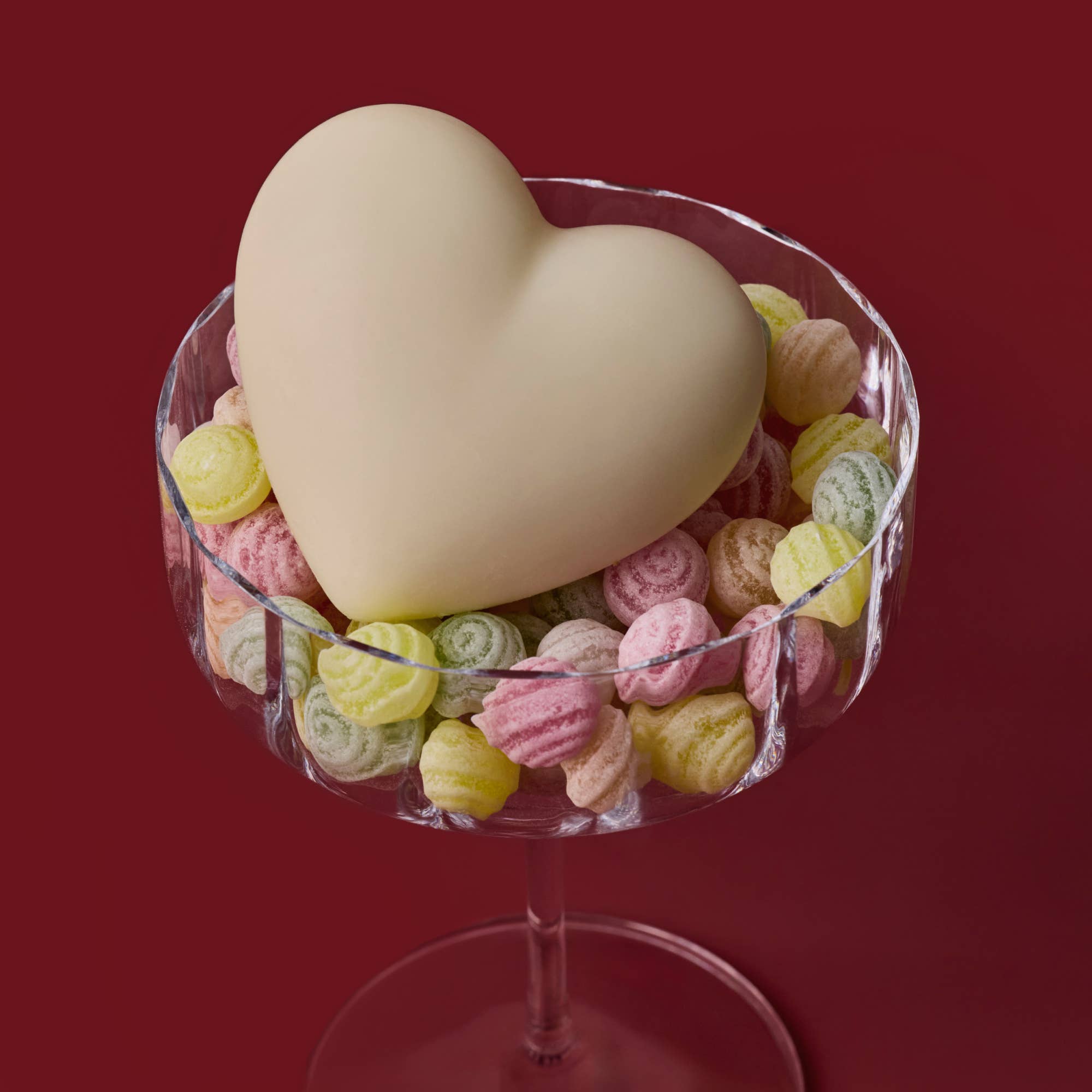 Heart-shaped white chocolate piece on a bed of colorful candy in a glass bowl against a red background