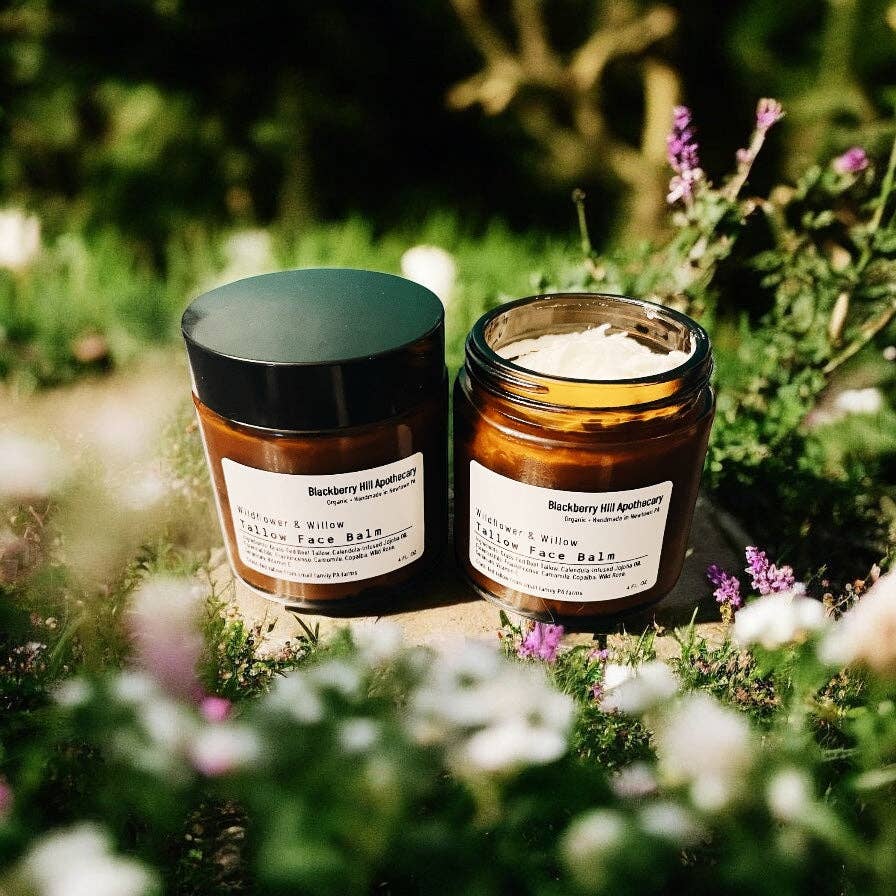 Two jars of 'Blackberry Hill Apothecary' balm on a natural background with greenery and flowers.