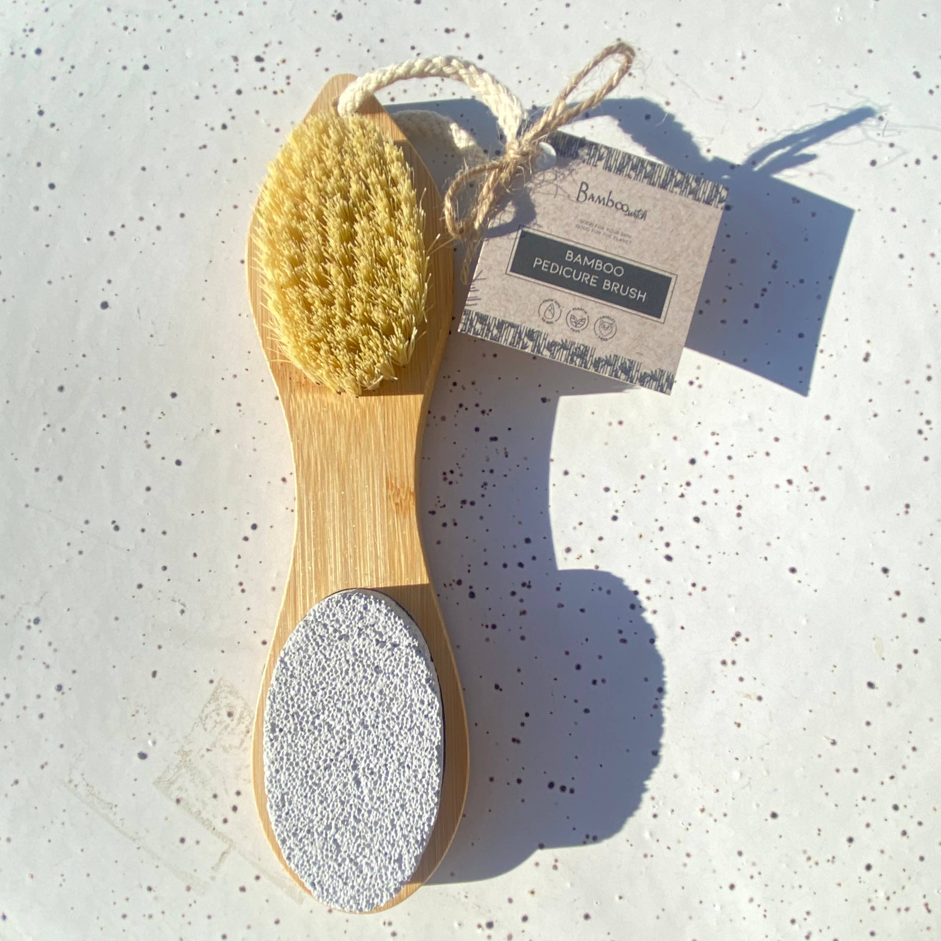 Bamboo pedicure brush with a gray scrubber and yellow bristle head on a speckled surface, accompanied by a branded tag.