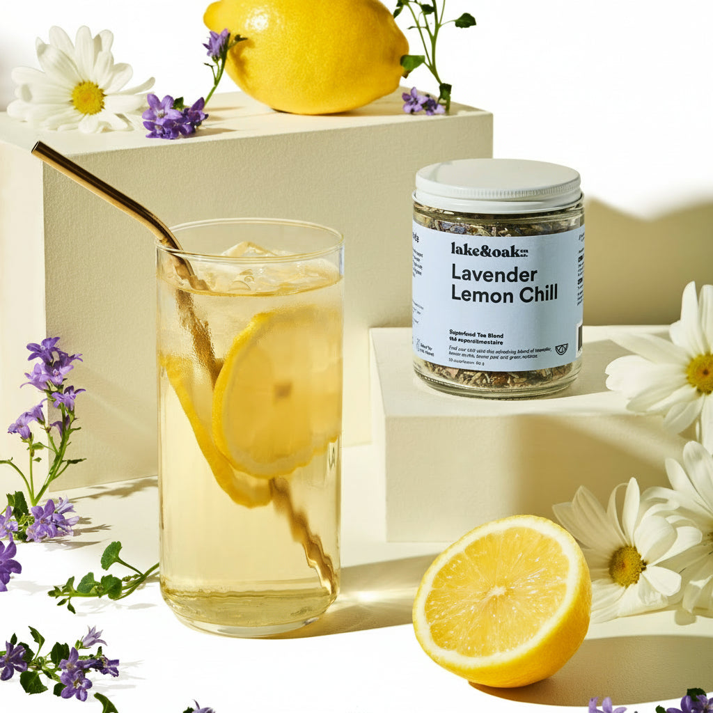 Glass of lemonade with a jar of 'Lavender Lemon Chill' tea blend, surrounded by lemons and flowers on a light background.